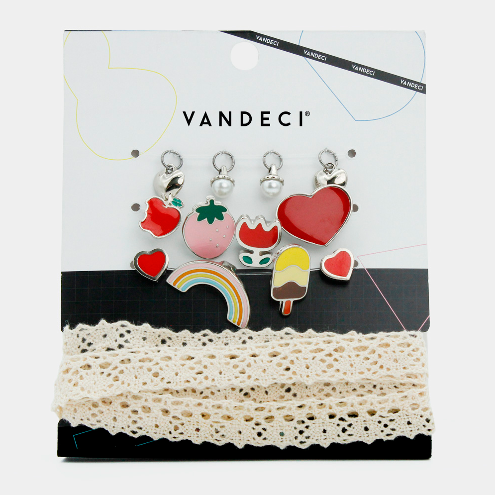 Set de charms Vandeci con figuras de corazón y fresa en su empaque original junto a agujetas de encaje beige.